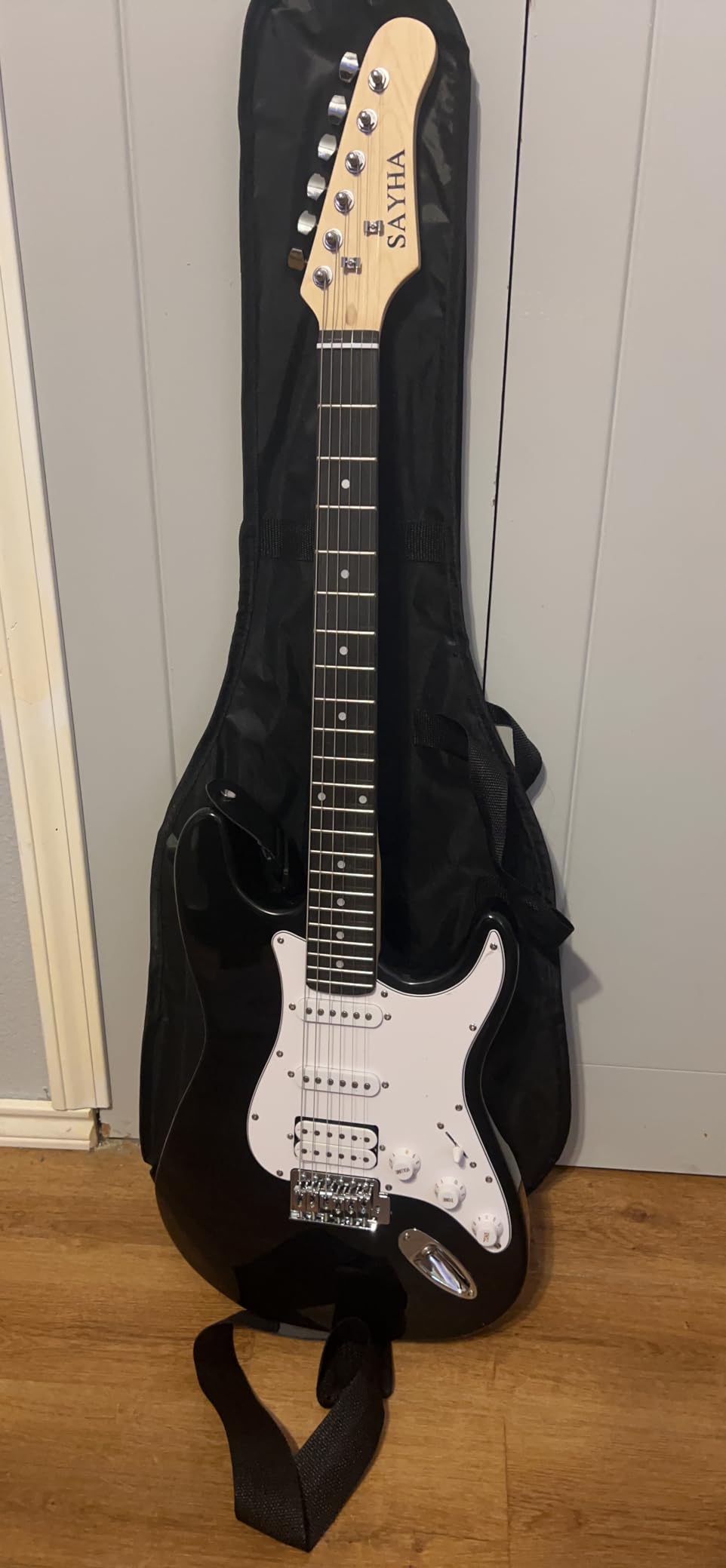 Black Sayha electric guitar with white pickguard standing in front of its gig bag, strap hanging below